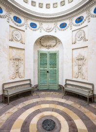 Schloss und Schlossgarten Schwetzingen - Garten - Tempel der Botanik - Von innen - Raum - Von Westen - Blick nach Osten in den Raum; in der Mitte die Tür des einzigen Zugangs zum Tempel; unten links und rechts zwei Stuckreliefs mit Dreifuß, Feuerschale und Schlange, links mit zwei gekreuzten Krummstäben mit Eichenlaub, den Wnter darstellend, rechts zwei gekreuzte Thyrsosstäbe (Bacchusstäbe) und Weinranken mit Trauben, den Herbst darstellend; oben links und rechts zwei stuckierte Reliefporträts von Naturforschern, links Plinius der Ältere (23-79), rechts Theophrastos von Eresos (371-287 v. Chr.);  unterhalb der Kuppel Girlanden, dazwischen Tierkreiszeichen und Stuckreliefs mit Putti, die Jahreszeiten darstellend; in der Kuppel rosettengeschmückte kleine Kassetten; auf dem Fußboden konzentrische Sandsteinplatten, ähnlich Jahresringen eines Baumes, in der Mitte eine kreisförmige Öffnung, bedeckt mit einem Blech, darunter ein dunkler, gewölbter, nicht zugänglicher Kellerraum (Tempel: Durchmesser ca. 7 Meter; Bauzeit: 1777-1780; Architekt: Nicolas de Pigage) (aufgenommen im September 2023, am frühen Nachmittag)