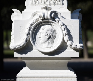 Schloss und Schlossgarten Schwetzingen - Garten - Kreisparterre, nordöstliches Rasenfeld, entlang der Nord-Süd-Achse - Nordöstlicher Obelisk, Ostseite - Blick auf ein männliches verwittertes Profilbildnis / Flachrelief, umgeben von einem Lorbeerfeston (Person nicht eindeutig identifizierbar) (Bildhauer: Peter Anton von Verschaffelt (1710-1793); Datierung: 1762-69; Obelisk Teil einer Vierergruppe im Kreisparterre; Material: roter Sandstein (Medaillons aus gelbem Sandstein); Fassung: Weiß; Obelisk dreigeteilt; Sockel, Block mit Medaillonbildnissen, eigentlicher Obelisk; Abmessungen: Höhe ca. 6 Meter, Sockel Höhe 155 cm / Breite 87 cm, Block mit Medaillons Höhe 67 cm / Breite 87 cm; Obelisk oben mit Tropfsteindekor) (aufgenommen im September 2023, am späten Vormittag)