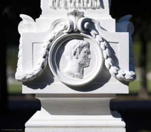 Schloss und Schlossgarten Schwetzingen - Garten - Kreisparterre, nordwestliches Rasenfeld, entlang der Nord-Süd-Achse - Nordwestlicher Obelisk, Ostseite - Blick auf ein männnliches verwittertes Profilbildnis / Flachrelief, umgeben von einem Lorbeerfeston (Person nicht eindeutig identifizierbar) (Bildhauer: Peter Anton von Verschaffelt (1710-1793); Datierung: 1762-69; Obelisk Teil einer Vierergruppe im Kreisparterre; Material: roter Sandstein (Medaillons aus gelbem Sandstein); Fassung: Weiß; Obelisk dreigeteilt; Sockel, Block mit Medaillonbildnissen, eigentlicher Obelisk; Abmessungen: Höhe ca. 6 Meter, Sockel Höhe 155 cm / Breite 87 cm, Block mit Medaillons Höhe 67 cm / Breite 87 cm; Obelisk oben mit Tropfsteindekor) (aufgenommen im September 2023, am späten Vormittag) Schloss und Schlossgarten Schwetzingen - Garten - Kreisparterre, nordwestliches Rasenfeld, entlang der Nord-Süd-Achse - Nordwestlicher Obelisk, Ostseite - Blick auf ein männnliches verwittertes Profilbildnis / Flachrelief, umgeben von einem Lorbeerfeston (Person nicht eindeutig identifizierbar) (Bildhauer: Peter Anton von Verschaffelt (1710-1793); Datierung: 1762-69; Obelisk Teil einer Vierergruppe im Kreisparterre; Material: roter Sandstein (Medaillons aus gelbem Sandstein); Fassung: Weiß; Obelisk dreigeteilt; Sockel, Block mit Medaillonbildnissen, eigentlicher Obelisk; Abmessungen: Höhe ca. 6 Meter, Sockel Höhe 155 cm / Breite 87 cm, Block mit Medaillons Höhe 67 cm / Breite 87 cm; Obelisk oben mit Tropfsteindekor) (aufgenommen im September 2023, am späten Vormittag)