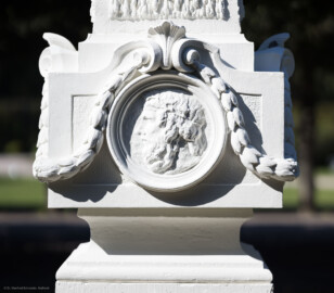 Schloss und Schlossgarten Schwetzingen - Garten - Kreisparterre, südwestliches Rasenfeld, entlang der Nord-Süd-Achse - Südwestlicher Obelisk, Ostseite - Blick auf ein männliches verwittertes Profilbildnis / Flachrelief, umgeben von einem Lorbeerfeston (Person nicht eindeutig identifizierbar) (Bildhauer: Peter Anton von Verschaffelt (1710-1793); Datierung: 1762-69; Obelisk Teil einer Vierergruppe im Kreisparterre; Material: roter Sandstein (Medaillons aus gelbem Sandstein); Fassung: Weiß; Obelisk dreigeteilt; Sockel, Block mit Medaillonbildnissen, eigentlicher Obelisk; Abmessungen: Höhe ca. 6 Meter, Sockel Höhe 155 cm / Breite 87 cm, Block mit Medaillons Höhe 67 cm / Breite 87 cm; Obelisk oben mit Tropfsteindekor) (aufgenommen im September 2023, am späten Vormittag)