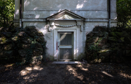 Schloss und Schlossgarten Schwetzingen - Garten - Minervatempel - Von aussen - Rückseite - Von Südwest - Blick auf den unteren Teil der Rückseite des Minervatempels mit den zwei Geschossen und (von unten nach oben) dem Portal zu einem unterirdischen Raum mit zwei Stufen, gerahmter Tür, Rosetten und Girlanden, Dreiecksgiebel mit Maske eines Pans bzw. Fauns, darüber die leere, verputzte Wand (Gesamter Bau: Breite ca. 7 Meter, Länge ca. 9 Meter, Innenraum kubisch; Rückseite zweigeschossig mit Kellerraum; Architekt: Nicolas de Pigage (1723-1796); Bauzeit: vermutlich zwischen 1767/68 und 1773) (aufgenommen im September 2023, am frühen Nachmittag)
