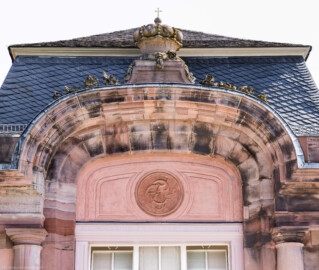 Schloss und Schlossgarten Schwetzingen - Garten - Nördlicher Zirkelbau - Westlicher Eck-Pavillon - Westlicher Abschluss - Blick nach oben auf die Bögen oberhalb der Fenster,  auf das Monogramm "CT" (Carl Theodor) sowie auf die Krone mit Kreuz  (Gesamter Bau: Länge Bogen ca. 175 Meter, Breite zwischen ca. 11 und 18 Meter, 51 Achsen, 5 Pavillons, 4 Zwischenbauten; Architekt: Alessandro Alessandro Galli da Bibiena (1686-1748), Bauleitung: zuerst Guillaume d’Hauberat (1680–1749), dann Nicolas de Pigage (1723-1796); Bauzeit: 1748-1750) (aufgenommen im September 2023, am frühen Nachmittag)