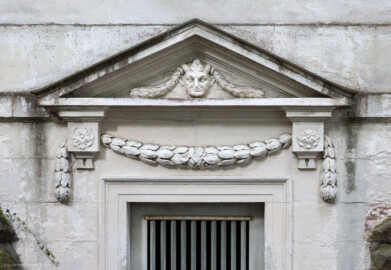 Schloss und Schlossgarten Schwetzingen - Garten - Minervatempel - Von aussen - Rückseite - Portal - Von Südwest - Blick auf einen Ausschnitt des Portals zu einem unterirdischen Raum, auf die Tür, den Türrahmen, Rosetten und Girlanden, den Dreiecksgiebel, darin eine Maske eines Pans bzw. Fauns (Gesamter Bau: Breite ca. 7 Meter, Länge ca. 9 Meter, Innenraum kubisch; Rückseite zweigeschossig mit Kellerraum; Architekt: Nicolas de Pigage (1723-1796); Bauzeit: vermutlich zwischen 1767/68 und 1773) (aufgenommen im September 2023, am Nachmittag)
