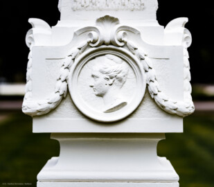 Schloss und Schlossgarten Schwetzingen - Garten - Kreisparterre, nordwestliches Rasenfeld, entlang der Nord-Süd-Achse - Nordwestlicher Obelisk, Nordseite - Blick auf ein weibliches verwittertes Profilbildnis / Flachrelief, umgeben von einem Lorbeerfeston (Person nicht eindeutig identifizierbar) (Bildhauer: Peter Anton von Verschaffelt (1710-1793); Datierung: 1762-69; Obelisk Teil einer Vierergruppe im Kreisparterre; Material: roter Sandstein (Medaillons aus gelbem Sandstein); Fassung: Weiß; Obelisk dreigeteilt; Sockel, Block mit Medaillonbildnissen, eigentlicher Obelisk; Abmessungen: Höhe ca. 6 Meter, Sockel Höhe 155 cm / Breite 87 cm, Block mit Medaillons Höhe 67 cm / Breite 87 cm; Obelisk oben mit Tropfsteindekor) (aufgenommen im September 2023, am späten Nachmittag)