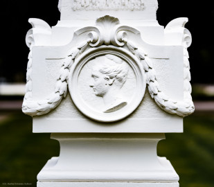 Schloss und Schlossgarten Schwetzingen - Garten - Kreisparterre, nordwestliches Rasenfeld, entlang der Nord-Süd-Achse - Nordwestlicher Obelisk, Nordseite - Blick auf ein weibliches verwittertes Profilbildnis / Flachrelief, umgeben von einem Lorbeerfeston (Person nicht eindeutig identifizierbar) (Bildhauer: Peter Anton von Verschaffelt (1710-1793); Datierung: 1762-69; Obelisk Teil einer Vierergruppe im Kreisparterre; Material: roter Sandstein (Medaillons aus gelbem Sandstein); Fassung: Weiß; Obelisk dreigeteilt; Sockel, Block mit Medaillonbildnissen, eigentlicher Obelisk; Abmessungen: Höhe ca. 6 Meter, Sockel Höhe 155 cm / Breite 87 cm, Block mit Medaillons Höhe 67 cm / Breite 87 cm; Obelisk oben mit Tropfsteindekor) (aufgenommen im September 2023, am späten Nachmittag) Schloss und Schlossgarten Schwetzingen - Garten - Kreisparterre, nordwestliches Rasenfeld, entlang der Nord-Süd-Achse - Nordwestlicher Obelisk, Nordseite - Blick auf ein weibliches verwittertes Profilbildnis / Flachrelief, umgeben von einem Lorbeerfeston (Person nicht eindeutig identifizierbar) (Bildhauer: Peter Anton von Verschaffelt (1710-1793); Datierung: 1762-69; Obelisk Teil einer Vierergruppe im Kreisparterre; Material: roter Sandstein (Medaillons aus gelbem Sandstein); Fassung: Weiß; Obelisk dreigeteilt; Sockel, Block mit Medaillonbildnissen, eigentlicher Obelisk; Abmessungen: Höhe ca. 6 Meter, Sockel Höhe 155 cm / Breite 87 cm, Block mit Medaillons Höhe 67 cm / Breite 87 cm; Obelisk oben mit Tropfsteindekor) (aufgenommen im September 2023, am späten Nachmittag)