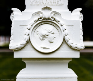 Schloss und Schlossgarten Schwetzingen - Garten - Kreisparterre, nordöstliches Rasenfeld, entlang der Nord-Süd-Achse - Nordöstlicher Obelisk, Nordseite - Blick auf ein weibliches verwittertes Profilbildnis / Flachrelief, umgeben von einem Lorbeerfeston (Person nicht eindeutig identifizierbar) (Bildhauer: Peter Anton von Verschaffelt (1710-1793); Datierung: 1762-69; Obelisk Teil einer Vierergruppe im Kreisparterre; Material: roter Sandstein (Medaillons aus gelbem Sandstein); Fassung: Weiß; Obelisk dreigeteilt; Sockel, Block mit Medaillonbildnissen, eigentlicher Obelisk; Abmessungen: Höhe ca. 6 Meter, Sockel Höhe 155 cm / Breite 87 cm, Block mit Medaillons Höhe 67 cm / Breite 87 cm; Obelisk oben mit Tropfsteindekor) (aufgenommen im September 2023, am späten Nachmittag)