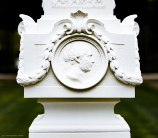 Schloss und Schlossgarten Schwetzingen - Garten - Kreisparterre, südöstliches Rasenfeld, entlang der Nord-Süd-Achse - Südöstlicher Obelisk, Nordseite - Blick auf ein weibliches verwittertes Profilbildnis / Flachrelief, umgeben von einem Lorbeerfeston (Person nicht eindeutig identifizierbar) (Bildhauer: Peter Anton von Verschaffelt (1710-1793); Datierung: 1762-69; Obelisk Teil einer Vierergruppe im Kreisparterre; Material: roter Sandstein (Medaillons aus gelbem Sandstein); Fassung: Weiß; Obelisk dreigeteilt; Sockel, Block mit Medaillonbildnissen, eigentlicher Obelisk; Abmessungen: Höhe ca. 6 Meter, Sockel Höhe 155 cm / Breite 87 cm, Block mit Medaillons Höhe 67 cm / Breite 87 cm; Obelisk oben mit Tropfsteindekor) (aufgenommen im September 2023, am späten Nachmittag)