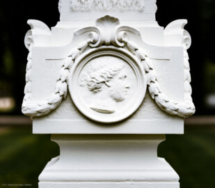 Schloss und Schlossgarten Schwetzingen - Garten - Kreisparterre, südwestliches Rasenfeld, entlang der Nord-Süd-Achse - Südwestlicher Obelisk, Nordseite - Blick auf ein weibliches verwittertes Profilbildnis / Flachrelief, umgeben von einem Lorbeerfeston (Person nicht eindeutig identifizierbar) (Bildhauer: Peter Anton von Verschaffelt (1710-1793); Datierung: 1762-69; Obelisk Teil einer Vierergruppe im Kreisparterre; Material: roter Sandstein (Medaillons aus gelbem Sandstein); Fassung: Weiß; Obelisk dreigeteilt; Sockel, Block mit Medaillonbildnissen, eigentlicher Obelisk; Abmessungen: Höhe ca. 6 Meter, Sockel Höhe 155 cm / Breite 87 cm, Block mit Medaillons Höhe 67 cm / Breite 87 cm; Obelisk oben mit Tropfsteindekor) (aufgenommen im September 2023, am späten Nachmittag)