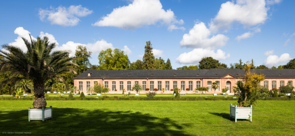 Schloss und Schlossgarten Schwetzingen - Garten - Neue Orangerie - Südseite - Blick vom südlichen Kanal des Orangeriegartens nach Norden auf einen Teil der Neuen Orangerie; links der westliche dreiachsige Eckpavillon, in der Mitte der westliche dreizehnachsige Seitenflügel, rechts der fünfachsiger Mittelbau, zweigeschossig, mit rundbogigem Portal, ganz rechts fünf Achsen des östlichen dreizehnachsigen Seitenflügels (Gesamter Bau: Länge ca. 171 Meter, Breite ca. 10-12 Meter; Bauzeit: 1761-1762; Architekt: Nicolas de Pigage; umfangreiche Instandsetzung: 1994-1999; Putzflächen bemalt in Freskotechnik, täuschen teilweise ein Scheinmauerwerk vor) (aufgenommen im Oktober 2023, um die Mittagszeit) Schloss und Schlossgarten Schwetzingen - Garten - Neue Orangerie - Südseite - Blick vom südlichen Kanal des Orangeriegartens nach Norden auf einen Teil der Neuen Orangerie; links der westliche dreiachsige Eckpavillon, in der Mitte der westliche dreizehnachsige Seitenflügel, rechts der fünfachsiger Mittelbau, zweigeschossig, mit rundbogigem Portal, ganz rechts fünf Achsen des östlichen dreizehnachsigen Seitenflügels (Gesamter Bau: Länge ca. 171 Meter, Breite ca. 10-12 Meter; Bauzeit: 1761-1762; Architekt: Nicolas de Pigage; umfangreiche Instandsetzung: 1994-1999; Putzflächen bemalt in Freskotechnik, täuschen teilweise ein Scheinmauerwerk vor) (aufgenommen im Oktober 2023, um die Mittagszeit)