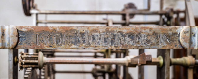 Schloss und Schlossgarten Schwetzingen - Schloss - Mittelbau - Von innen - Dachstuhl, Südseite - Uhrenstube im südlichen Turm - Von Osten - Blick auf die Inschrift "IACOB MOELLINGER" am rechten / östlichen oberen Rahmen der großen alten Schlossuhr von Johann Jacob Möllinger (1695-1763) mit Viertel- und Stundenschlag, stillgelegt 1968; die gesamte umlaufende Inschrift am oberen Rahmen der Turmuhr lautet: "AUF HOHEN CHURFÜRSTLICHEN BEFEHL WURDE DIESES WERCK VON IACOB MOELLINGER IN NEUSTATT VERFERTIGET Ao. 1763" (Mittelbau: Länge Westseite ca. 45 Meter, Breite Süd-/Nordseite ca. 38 Meter; Wiederaufbau / Erweiterung: 1698-1717; Architekten: Johann Adam Breunig, Simon Sarto) (aufgenommen im November 2023, am Nachmittag) Schloss und Schlossgarten Schwetzingen - Schloss - Mittelbau - Von innen - Dachstuhl, Südseite - Uhrenstube im südlichen Turm - Von Osten - Blick auf die Inschrift "IACOB MOELLINGER" am rechten / östlichen oberen Rahmen der großen alten Schlossuhr von Johann Jacob Möllinger (1695-1763) mit Viertel- und Stundenschlag, stillgelegt 1968; die gesamte umlaufende Inschrift am oberen Rahmen der Turmuhr lautet: "AUF HOHEN CHURFÜRSTLICHEN BEFEHL WURDE DIESES WERCK VON IACOB MOELLINGER IN NEUSTATT VERFERTIGET Ao. 1763" (Mittelbau: Länge Westseite ca. 45 Meter, Breite Süd-/Nordseite ca. 38 Meter; Wiederaufbau / Erweiterung: 1698-1717; Architekten: Johann Adam Breunig, Simon Sarto) (aufgenommen im November 2023, am Nachmittag)