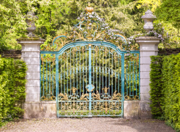 Schloss und Schlossgarten Schwetzingen - Garten - Arboretum - Südliches Tor - Von Süden - Blick auf das schmiedeeiserne, teilvergoldete Prachttor, der südliche Eingang zum Arboretum, mit Rokoko-Verzierungen, oben das Monogramm "CT" (Carl Theodor) und ganz oben die Krone (Eisentür fertiggestellt 1755; entworfen von Franz Wilhelm Rabaliatti, geschmiedet von den Mannheimer Schlossermeistern Strickling und Frockmann; ursprünglich an einem anderen Ort verbaut) (aufgenommen im April 2024, am frühen Nachmittag)