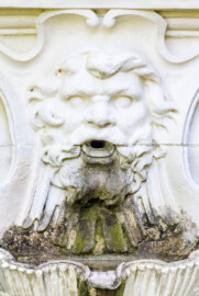 Schloss und Schlossgarten Schwetzingen - Garten - Naturtheater -  Querachse, südliches Ende, an Gartenmauer - Delphinbrunnen / "fontaine du mascaron" - Von Norden - Blick auf die bärtige Pansmaske als Wasserspender des Delphinbrunnens; Wandbrunnen, gespeist aus Überlaufwasser des Auffangbeckens der Kaskade beim Apollotempel (Bildhauer: Peter Anton von Verschaffelt (1710-1793), Datierung: vor 1775; Brunnen: Material gelber Sandstein, weiß gefasst, Höhe ca. 2,4 Meter, Beckenbreite ca. 2,5 Meter)  (aufgenommen im April 2024, am Nachmittag)