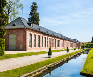Schloss und Schlossgarten Schwetzingen - Garten - Neue Orangerie - West- und Südseite - Von Südwest - Blick vom westlichen Kanal des Orangeriegartens auf die Westseite (links) und auf die Südseite (Mitte) der Neuen Orangerie; Aufbau der Orangerie: fünfachsiger Mittelbau, zweigeschossig, mit rundbogigem Portal; links und rechts davon dreizehnachsige Seitenflügel, jeweils in der Mitte eine Tür; links und rechts der Seitenflügel dreiachsige Eckpavillons; ganz rechts ein nachträglich angebautes Glashaus; ein geplantes Glashaus ganz links ist nicht realisiert (Gesamter Bau: Länge ca. 171 Meter, Breite ca. 10-12 Meter; Bauzeit: 1761-1762, Glashaus angebaut 1770; Architekt: Nicolas de Pigage; umfangreiche Instandsetzung: 1994-1999) (aufgenommen im April 2024, um die Mittagszeit)