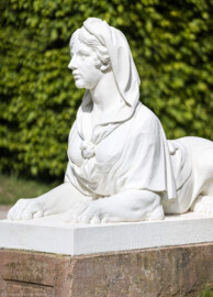 Schloss und Schlossgarten Schwetzingen - Garten - Naturtheater - Parterre - Ostseite, südliche Sphynx - Von Südwest - Blick auf eine der sechs Sphyngen (als Allegorien der Musik, des Tanzes, der Komödie, der Tragödie und als Wächterinnen) neben der Treppe zum Zuschauerraum, auf einem Postament liegend (Bildhauer: Peter Anton von Verschaffelt (1710-1793), Datierung: 1766-1773; Material: gelber Sandstein, weiß gefasst, Höhe ca. 100 cm, Postament 160 x 63 cm) (aufgenommen im April 2024, am Nachmittag)