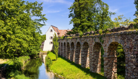 Schloss und Schlossgarten Schwetzingen - Sonstige Bauten - Unteres Wasserwerk - Von Süden - Blick vom westlichen Aquädukt direkt neben dem Römischen Wasserkastell nach Norden auf das Untere Wasserwerk, rechts der nördliche Aquädukt zum Wasserwerk, unten mittig der Leimbach nach Verlassen des Schlossgartens in Fließrichtung Wasserwerk bzw. Norden  (Gesamter Bau: Länge x Breite x Höhe: ca. 25 x 13 x 18  Meter; Wasserturm (mit Radkammer, 2 Wasserreservoirs, 3 Geschosse): ca. 13 x 8 Meter, Pumpenhaus (mit Pumpenstube, 2 Geschosse): ca. 14 x 13 Meter, Knochenmühle: ca. 8 x 6 Meter; Architekt: Nicolas de Pigage (1723-1796); Bauzeit: ca. 1762-1765) (aufgenommen im Mai 2024, am frühen Nachmittag)