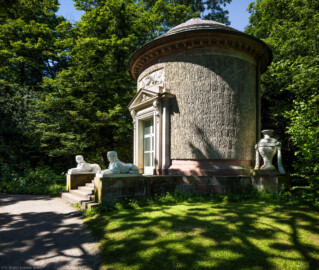 Schloss und Schlossgarten Schwetzingen - Garten - Tempel der Botanik - Von außen - Von Nordost - Blick auf den Tempel der Botanik; ein zylindrischer Rundtempel, einem Baumstumpf mit Eichenrinde ähnlich, mit siebenstufiger Treppe, zwei Sphinxen, einem Portal mit Holzlamellen-Flügeltür, Dreiecksgiebel, darin ein geflügelter weiblicher Kopf, darunter eine Inschrift, darüber ein Relief mit zwei Greifen, darüber eine flache Laterne, seitlich zwei steinerne Urnen (Sphinxen Peter Simon Lamine (1738-1817) zugeschrieben; Portal von Franz Conrad Linck (1730-1793); seitliche Urnen von Johann Matthäus van den Branden (1716-1788); Inschrift "Botanicae Silvestri Anno MDCCLXXVIII", deutsch: "Der Waldbotanik im Jahre 1778") (Tempel: Durchmesser ca. 7 Meter; Bauzeit: 1777-1780; Architekt: Nicolas de Pigage) (aufgenommen im Mai 2024, am späten Vormittag)
