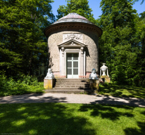 Schloss und Schlossgarten Schwetzingen - Garten - Tempel der Botanik - Von außen - Von Osten - Blick auf den Tempel der Botanik; ein zylindrischer Rundtempel, einem Baumstumpf mit Eichenrinde ähnlich, mit siebenstufiger Treppe, zwei Sphinxen, einem Portal mit Holzlamellen-Flügeltür, Dreiecksgiebel, darin ein geflügelter weiblicher Kopf, darunter eine Inschrift, darüber ein Relief mit zwei Greifen, darüber eine flache Laterne, seitlich zwei steinerne Urnen (Sphinxen Peter Simon Lamine (1738-1817) zugeschrieben; Portal von Franz Conrad Linck (1730-1793); seitliche Urnen von Johann Matthäus van den Branden (1716-1788); Inschrift "Botanicae Silvestri Anno MDCCLXXVIII", deutsch: "Der Waldbotanik im Jahre 1778") (Tempel: Durchmesser ca. 7 Meter; Bauzeit: 1777-1780; Architekt: Nicolas de Pigage) (aufgenommen im Mai 2024, um die Mittagszeit)
