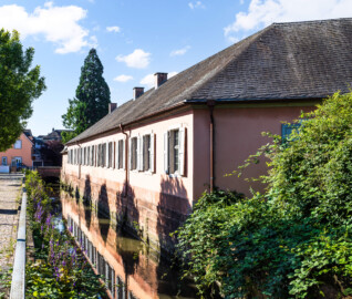 Schloss und Schlossgarten Schwetzingen - Garten - Invalidenkaserne (ehemaliges Baumagazin) / Stadtbibliothek  - Ostflügel - Von aussen - Von Nordost - Blick nach Südost auf die Fassade des Ostflügels; im Inneren des Flügels befindet sich die Stadtbibliothek; der Kanal ist Teil des Leimbachs und fliesst westlich Richtung Unteres Wasserwerk (Vierflügelanlage: Länge x Breite: ca. 60 x 50 Meter; Ostflügel: ca. 60 x 10 Meter; Bauzeit: um 1774; Architekt des Baummagazins: Nicolas de Pigage; Umbau zur Kaserne: Ingenieur-Oberst von Pfister) (aufgenommen im Juni 2024, am Vormittag)