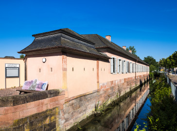 Schloss und Schlossgarten Schwetzingen - Garten - Invalidenkaserne (ehemaliges Baumagazin) / Stadtbibliothek - Ostflügel - Von aussen - Von Südost - Blick nach Nordwest auf die Fassade des Ostflügels; im Inneren des Flügels befindet sich die Stadtbibliothek; links der Eingang zur Bibliothek; der Kanal rechts ist Teil des Leimbachs und fliesst westlich Richtung Unteres Wasserwerk (Vierflügelanlage: Länge x Breite: ca. 60 x 50 Meter; Ostflügel: ca. 60 x 10 Meter; Bauzeit: um 1774; Architekt des Baummagazins: Nicolas de Pigage; Umbau zur Kaserne: Ingenieur-Oberst von Pfister) (aufgenommen im Juni 2024, am Vormittag) Schloss und Schlossgarten Schwetzingen - Garten - Invalidenkaserne (ehemaliges Baumagazin) / Stadtbibliothek - Ostflügel - Von aussen - Von Südost - Blick nach Nordwest auf die Fassade des Ostflügels; im Inneren des Flügels befindet sich die Stadtbibliothek; links der Eingang zur Bibliothek; der Kanal rechts ist Teil des Leimbachs und fliesst westlich Richtung Unteres Wasserwerk (Vierflügelanlage: Länge x Breite: ca. 60 x 50 Meter; Ostflügel: ca. 60 x 10 Meter; Bauzeit: um 1774; Architekt des Baummagazins: Nicolas de Pigage; Umbau zur Kaserne: Ingenieur-Oberst von Pfister) (aufgenommen im Juni 2024, am Vormittag)