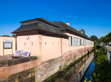 Schloss und Schlossgarten Schwetzingen - Garten - Invalidenkaserne (ehemaliges Baumagazin) / Stadtbibliothek  - Ostflügel - Von aussen - Von Südost - Blick nach Nordwest auf die Fassade des Ostflügels; im Inneren des Flügels befindet sich die Stadtbibliothek; links der Eingang zur Bibliothek; der Kanal rechts ist Teil des Leimbachs und fliesst westlich Richtung Unteres Wasserwerk (Vierflügelanlage: Länge x Breite: ca. 60 x 50 Meter; Ostflügel: ca. 60 x 10 Meter; Bauzeit: um 1774; Architekt des Baummagazins: Nicolas de Pigage; Umbau zur Kaserne: Ingenieur-Oberst von Pfister) (aufgenommen im Juni 2024, am Vormittag)