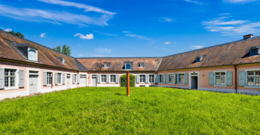 Schloss und Schlossgarten Schwetzingen - Garten - Invalidenkaserne (ehemaliges Baumagazin) - Innenhof - Von Süden - Blick von der Tordurchfahrt im Südflügel nach Norden in den abgeschlossenen Innenhof; links der Westflügel (genutzt als Ausstellungsraum von xylon), in der Mitte der Nordflügel (genutzt als Werkstattsraum von xylon) mit der durch Laub verdeckten nördlichen Tordurchfahrt , rechts der Ostflügel (genutzt von der Stadtbibliothek) (Vierflügelanlage: Länge x Breite: ca. 60 x 50 Meter; Südflügel: ca. 60 x 10 Meter; Bauzeit: um 1774; Architekt des Baummagazins: Nicolas de Pigage; Umbau zur Kaserne: Ingenieur-Oberst von Pfister) (aufgenommen im Juni 2024, am späten Vormittag)