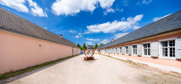 Schloss und Schlossgarten Schwetzingen - Garten - Invalidenkaserne (ehemaliges Baumagazin) / Baumagazin - Außenhof - Von Osten - Blick nach Westen in den abschließbaren Außenhof zwischen dem Baumagazin (links, mit der Gartengeräte-Ausstellung) und dem Südflügel der Invalidenkaserne (rechts); im Hintergrund zwischen den Bäumen die Ostseite des Glashauses der Neuen Orangerie; die vier Fenster rechts der Mitte gehören zu xylon, die sieben Fenster ganz rechts gehören zur Stadtbibliothek (Vierflügelanlage: Länge x Breite: ca. 60 x 50 Meter; Südflügel: ca. 60 x 10 Meter; Bauzeit: um 1774; Architekt des Baummagazins: Nicolas de Pigage; Umbau zur Kaserne: Ingenieur-Oberst von Pfister) (aufgenommen im Juni 2024, um die Mittagszeit)