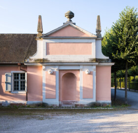 Schloss und Schlossgarten Schwetzingen - Garten - Invalidenkaserne (ehemaliges Baumagazin) - Nordflügel, Westseite - Fontainenportal - Von Norden - Blick auf das sog. Fontainenportal, eine Giebelfassade mit Ziergliedern aus Sandstein und verputzten Wandflächen; ganz oben eine Deckelvase, links und rechts ein Obelisk, dazwischen ein Giebelfeld, darunter ein (heute leeres) Rechteckfeld (ursprünglich mit dem kurfürstlichen Wappen geschmückt), darunter das Kranzgesims, darunter drei Felder, mittig mit Rundbogennische (ehemals versehen mit einer Muschel); unterhalb des Gesims vier quadratische Flächen, darin runde Scheiben, darunter je drei Guttae; oben in den äußeren beiden Wandfeldern zwei in die Horizontale gekippte und abgeschnittene wasserspendenden Vasen; Wandflächen ursprünglich mit einer tropfsteinartigen Oberfläche versehen (Vierflügelanlage: Länge x Breite: ca. 60 x 50 Meter; Nordflügel: ca. 30 x 10 Meter; Bauzeit: um 1774; Architekt des Baummagazins: Nicolas de Pigage; Umbau zur Kaserne: Ingenieur-Oberst von Pfister) (aufgenommen im Juli 2024, am frühen Vormittag)
