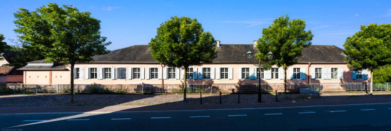 Schloss und Schlossgarten Schwetzingen - Garten - Invalidenkaserne (ehemaliges Baumagazin) / Stadtbibliothek  - Ostflügel - Von aussen - Von Nordost - Blick von der Turnhalle der Hildaschule an der Kronenstraße auf die gesamte Fassade des Ostflügels; Ostflügel genutzt von der Stadtbibliothek; hinter/unterhalb des Geländers fliesst der Leimbachkanal (Vierflügelanlage: Länge x Breite: ca. 60 x 50 Meter; Ostflügel: ca. 60 x 10 Meter; Bauzeit: um 1774; Architekt des Baummagazins: Nicolas de Pigage; Umbau zur Kaserne: Ingenieur-Oberst von Pfister) (aufgenommen im Juli 2024, am Vormittag)