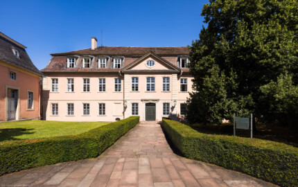 Schloss und Schlossgarten Schwetzingen - Garten - Gesandtenhaus (ehemaliges Prinzenhaus) / Amtsgericht - Von Südost - Blick von der Zeyherstraße auf die Südostfassade und das Portal des Gesandtenhauses; links die Rückseite des mittleren Pavillons des nördlichen Zirkelbaus; der Baum rechts ist eine Sommer-Linde (Tilia platyphyllos, Baum-Nr. 69)  (Nutzung: Wohnungen für Angehörige der kurfürstlichen Familie, ausländische Besucher, Nicolas de Pigage (1723-1796), Friedrich Ludwig Skell (1750-1823), Johann Michael Zeyher (1770-1843), Sterbeort von Johann Peter Hebel (1760-1826), ab 1805 Amtsgericht) (Bau: Länge x Breite: ca. 40 x 17 Meter; Bauzeit: um 1723; Architekt: unbekannt) (aufgenommen im Juli 2024, am späten Vormittag)