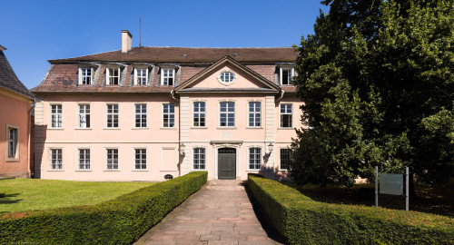 Schloss und Schlossgarten Schwetzingen - Garten - Gesandtenhaus (ehemaliges Prinzenhaus) / Amtsgericht - Von Südost - Blick von der Zeyherstraße auf die Südostfassade und das Portal des Gesandtenhauses; links die Rückseite des mittleren Pavillons des nördlichen Zirkelbaus; der Baum rechts ist eine Sommer-Linde (Tilia platyphyllos, Baum-Nr. 69) (Nutzung: Wohnungen für Angehörige der kurfürstlichen Familie, ausländische Besucher, Nicolas de Pigage (1723-1796), Friedrich Ludwig Skell (1750-1823), Johann Michael Zeyher (1770-1843), Sterbeort von Johann Peter Hebel (1760-1826), ab 1805 Amtsgericht) (Bau: Länge x Breite: ca. 40 x 17 Meter; Bauzeit: um 1723; Architekt: unbekannt) (aufgenommen im Juli 2024, am späten Vormittag) Schloss und Schlossgarten Schwetzingen - Garten - Gesandtenhaus (ehemaliges Prinzenhaus) / Amtsgericht - Von Südost - Blick von der Zeyherstraße auf die Südostfassade und das Portal des Gesandtenhauses; links die Rückseite des mittleren Pavillons des nördlichen Zirkelbaus; der Baum rechts ist eine Sommer-Linde (Tilia platyphyllos, Baum-Nr. 69) (Nutzung: Wohnungen für Angehörige der kurfürstlichen Familie, ausländische Besucher, Nicolas de Pigage (1723-1796), Friedrich Ludwig Skell (1750-1823), Johann Michael Zeyher (1770-1843), Sterbeort von Johann Peter Hebel (1760-1826), ab 1805 Amtsgericht) (Bau: Länge x Breite: ca. 40 x 17 Meter; Bauzeit: um 1723; Architekt: unbekannt) (aufgenommen im Juli 2024, am späten Vormittag)