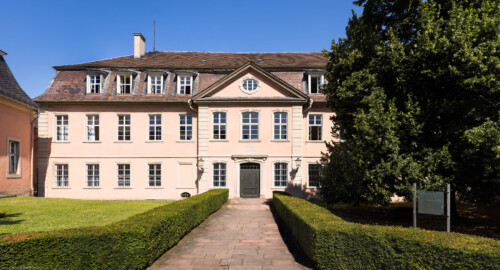 Schloss und Schlossgarten Schwetzingen - Garten - Gesandtenhaus (ehemaliges Prinzenhaus) / Amtsgericht - Von Südost - Blick von der Zeyherstraße auf die Südostfassade und das Portal des Gesandtenhauses; links die Rückseite des mittleren Pavillons des nördlichen Zirkelbaus; der Baum rechts ist eine Sommer-Linde (Tilia platyphyllos, Baum-Nr. 69) (Nutzung: Wohnungen für Angehörige der kurfürstlichen Familie, ausländische Besucher, Nicolas de Pigage (1723-1796), Friedrich Ludwig Skell (1750-1823), Johann Michael Zeyher (1770-1843), Sterbeort von Johann Peter Hebel (1760-1826), ab 1805 Amtsgericht) (Bau: Länge x Breite: ca. 40 x 17 Meter; Bauzeit: um 1723; Architekt: unbekannt) (aufgenommen im Juli 2024, am späten Vormittag) Schloss und Schlossgarten Schwetzingen - Garten - Gesandtenhaus (ehemaliges Prinzenhaus) / Amtsgericht - Von Südost - Blick von der Zeyherstraße auf die Südostfassade und das Portal des Gesandtenhauses; links die Rückseite des mittleren Pavillons des nördlichen Zirkelbaus; der Baum rechts ist eine Sommer-Linde (Tilia platyphyllos, Baum-Nr. 69) (Nutzung: Wohnungen für Angehörige der kurfürstlichen Familie, ausländische Besucher, Nicolas de Pigage (1723-1796), Friedrich Ludwig Skell (1750-1823), Johann Michael Zeyher (1770-1843), Sterbeort von Johann Peter Hebel (1760-1826), ab 1805 Amtsgericht) (Bau: Länge x Breite: ca. 40 x 17 Meter; Bauzeit: um 1723; Architekt: unbekannt) (aufgenommen im Juli 2024, am späten Vormittag)