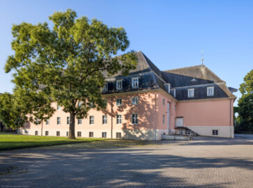 Schloss und Schlossgarten Schwetzingen - Garten - Schlosstheater - Von Nordwest - Blick vom Parkplatz des Theaters (Teil des Gesandtenhausgartens) nach Süden auf die Nordfassade (links) und die Westfassade (rechts) des Theaterbaus; die rechten 7 Fenster der Nordfassade gehören zur Seitenbühne; hinter der Wand links davon befindet sich das Foyer und Treppenhaus, dahinter der Zuschauerrraum; hinter der großen Wand rechts befindet sich die Hinterbühne; links davor eine flache Auffahrt zur Rampe und dem großen Tor der Seitenbühne; dahinter ein (Not-) Ausgang der Hinterbühne; dazwischen (kaum sichtbar) eine Treppe nach unten zum nördlichen Eingang in das (etwas tiefer liegende) Erdgeschoss; links ein großer Schnurbaum (Sophora japonica, Baum-Nr. 85) (Gesamter Bau: Länge x Breite: ca. 62 x 35 Meter; Architekt: Nicolas de Pigage (1723-1796); Bauzeit: 1752-1753, Erweiterung 1762, Sanierung: 1937, Renovierung: 1952, Generalsanierung / Umbau 1971-1974, Restaurierung: 1999-2005) (aufgenommen im Juli 2024, am frühen Vormittag)