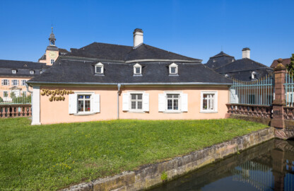 Schloss und Schlossgarten Schwetzingen - Schloss - Ehrenhof / Cour d'honneur - Südliches Wachthaus / Kurfürstenstube - Von Südost - Blick von der Schlossstraße südlich des Eingangs zum Ehrenhof auf die Ostseite des ehemaligen südlichen Wachthauses, heute als Café ("Kurfürstenstube") genutzt; im Vordergrund der Leimbach, im Hintergrund ganz links der Querbau des südlichen Flügels / des Hofdamenflügels (heute von der Justizakademie genutzt), rechts oberhalb davon der südliche Treppenturm und der südliche Turm, ganz rechts die Brücke über den Leimbach, links davon (hinter dem Gitter) das nördliche Wachthaus (heute als Kasse und Shop genutzt), links oberhalb davon das Dach des oberen Wasserwerks (aufgenommen im Juli 2024, am späten Vormittag)
