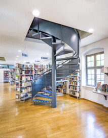 Schloss und Schlossgarten Schwetzingen - Garten - Invalidenkaserne (ehemaliges Baumagazin) / Stadtbibliothek  - Ostflügel - Von innen - Erdgeschoss - Von Südost - Blick nach Nordwest in den Bibliotheksraum und auf die Spindeltreppe zum Dachgeschoss (Vierflügelanlage: Länge x Breite: ca. 60 x 50 Meter; Ostflügel: ca. 60 x 10 Meter; Bauzeit: um 1774; Architekt des Baummagazins: Nicolas de Pigage; Umbau zur Kaserne: Ingenieur-Oberst von Pfister) (aufgenommen im Juli 2024, am frühen Nachmittag)