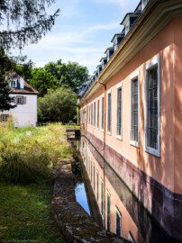 Schloss und Schlossgarten Schwetzingen - Schloss - Küchenflügel / Küchenbau - Längsbau, Südseite - Von Südost - Blick vom östlichen Ende des Längsbaus nach Westen auf die Südfassade des Längsbaus, auf den Leimbach-Kanal, die kleine Brücke zum südlichen Eingang zum Längsbau sowie auf das Studentenhaus (links) (Küchenflügel: L-förmig, Länge x Breite Längsbau ca. 75 x 13 Meter, Querbau ca. 25 x 11 Meter; Architekt: Nicolas de Pigage (1723-1796); Fertigstellung: 1761, letzte Renovierung: 2009-2012) (aufgenommen im August 2024, am frühen Nachmittag)