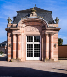 Schloss und Schlossgarten Schwetzingen - Garten - Nördlicher Zirkelbau - Östlicher Eck-Pavillon - Von Süden - Blick auf die Südfassade des Pavillons mit der großen Tür; oben das Monogramm "CT" (Carl Theodor), die Krone mit Kreuz sowie Blumenkörbe (Gesamter Bau: Länge Bogen ca. 175 Meter, Breite zwischen ca. 11 und 18 Meter, 51 Achsen, 5 Pavillons, 4 Zwischenbauten; Architekt: Alessandro Alessandro Galli da Bibiena (1686-1748), Bauleitung: zuerst Guillaume d’Hauberat (1680–1749), dann Nicolas de Pigage (1723-1796); Bauzeit: 1748-1750) (aufgenommen im August 2024, am Vormittag)