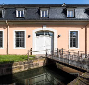 Schloss und Schlossgarten Schwetzingen - Schloss - Küchenflügel / Küchenbau - Längsbau, Südseite - Von Süden - Blick von der Nordwand des Studentenhauses auf die Brücke über den Leimbachkanal zum Eingang sowie auf das Tor zum Küchenbau (Küchenflügel: L-förmig, Länge x Breite Längsbau ca. 75 x 13 Meter, Querbau ca. 25 x 11 Meter; Architekt: Nicolas de Pigage (1723-1796); Fertigstellung: 1761, letzte Renovierung: 2009-2012) (aufgenommen im August 2024, am späten Vormittag)
