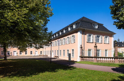 Schloss und Schlossgarten Schwetzingen - Schloss - Hof zwischen Südflügel und Küchenflügel - Von Südost - Blick von der Rasenfläche des Hofs nach Nordwest auf die Ostfassade (rechts) und die Südfassade (Mitte) des Südflügels sowie auf die Ostfassade des Querbaus des Küchenflügels (links); ganz rechts ein Ende des Gitterzauns um den Ehrenhof des Schlosses; die vier Bäume des Innenhofs sind Holländische Linden (Tilia × europaea) (Südflügel: L-förmig, Länge x Breite Längsbau ca. 50 x 14 Meter, Querbau ca. 15 x 11 Meter; Architekt: Johann Adam Breunig (1660-1727); Fertigstellung: 1710, letzte Renovierung: 2009-2012) (aufgenommen im August 2024, um die Mittagszeit)
