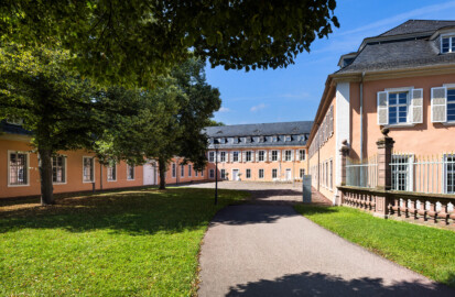 Schloss und Schlossgarten Schwetzingen - Schloss - Hof zwischen Südflügel und Küchenflügel - Von Osten - Blick vom Eingang nach Westen in den Hof, auf die Ostfassade des Südflügels (rechts), auf die Ostfassade des Querbaus des Küchenflügels (Mitte) sowie auf die Nordfassade des Längsbaus des Küchenbaus (links); ganz rechts ein Ende des Gitterzauns um den Ehrenhof des Schlosses; die vier Bäume des Innenhofs sind Holländische Linden (Tilia × europaea) (Südflügel: L-förmig, Länge x Breite Längsbau ca. 50 x 14 Meter, Querbau ca. 15 x 11 Meter; Architekt: Johann Adam Breunig (1660-1727); Fertigstellung: 1710; Küchenflügel: L-förmig, Länge x Breite Längsbau ca. 75 x 13 Meter, Querbau ca. 25 x 11 Meter; Architekt: Nicolas de Pigage (1723-1796); Fertigstellung: 1761; letzte Renovierung beider Flügel: 2009-2012) (aufgenommen im August 2024, um die Mittagszeit)
