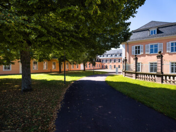 Schloss und Schlossgarten Schwetzingen - Schloss - Hof zwischen Südflügel und Küchenflügel - Von Osten - Blick vom Eingang nach Westen in den Hof, auf die Ostfassade des Südflügels (rechts), auf die Ostfassade des Querbaus des Küchenflügels (Mitte) sowie auf die Nordfassade des Längsbaus des Küchenbaus (links); ganz rechts ein Ende des Gitterzauns um den Ehrenhof des Schlosses; die vier Bäume des Innenhofs sind Holländische Linden (Tilia × europaea) (Südflügel: L-förmig, Länge x Breite Längsbau ca. 50 x 14 Meter, Querbau ca. 15 x 11 Meter; Architekt: Johann Adam Breunig (1660-1727); Fertigstellung: 1710; Küchenflügel: L-förmig, Länge x Breite Längsbau ca. 75 x 13 Meter, Querbau ca. 25 x 11 Meter; Architekt: Nicolas de Pigage (1723-1796); Fertigstellung: 1761; letzte Renovierung beider Flügel: 2009-2012) (aufgenommen im August 2024, am frühen Nachmittag)