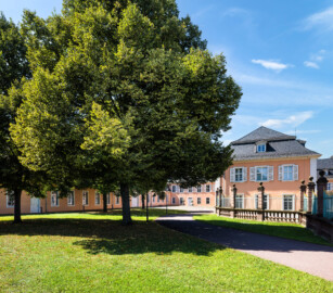 Schloss und Schlossgarten Schwetzingen - Schloss - Hof zwischen Südflügel und Küchenflügel - Von Osten - Blick vom Eingang nach Westen in den Hof, auf die Ostfassade des Südflügels (rechts), auf die Ostfassade des Querbaus des Küchenflügels (Mitte) sowie auf die Nordfassade des Längsbaus des Küchenbaus (links); ganz rechts ein Ende des Gitterzauns um den Ehrenhof des Schlosses; die vier Bäume des Innenhofs sind Holländische Linden (Tilia × europaea) (Südflügel: L-förmig, Länge x Breite Längsbau ca. 50 x 14 Meter, Querbau ca. 15 x 11 Meter; Architekt: Johann Adam Breunig (1660-1727); Fertigstellung: 1710; Küchenflügel: L-förmig, Länge x Breite Längsbau ca. 75 x 13 Meter, Querbau ca. 25 x 11 Meter; Architekt: Nicolas de Pigage (1723-1796); Fertigstellung: 1761; letzte Renovierung beider Flügel: 2009-2012) (aufgenommen im August 2024, am frühen Nachmittag)