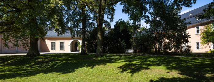 Schloss und Schlossgarten Schwetzingen - Schloss - Verbindungsbau Küchenbau-Zirkelbau / Alte Galerie - Südseite - Von Süden - Blick vom Seepferdgarten nach Norden auf die Südseite des Verbindungsbaus; ganz links die Ostseite des südlichen Zirkelbaus, rechts davon der Durchgang vom Seepferdgarten zur Schlossterrasse, in der Mitte (verdeckt durch Bäume und Sträucher) der östliche Teil der Südfassade, ganz rechts die Westseite des Querbaus des Küchenflügels (Verbindungsbau: Länge ca. 45 Meter, Breite ca. 8 Meter; Bauzeit: wohl um 1724; Architekt: unklar, evtl. Alessandro Galli da Bibiena (1686-1748) oder Johann Adam Breunig (1660-1727)) (aufgenommen im August 2024, am Nachmittag)