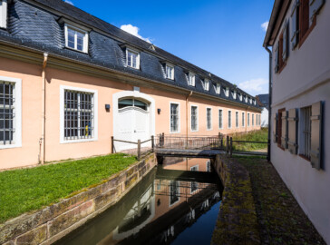 Schloss und Schlossgarten Schwetzingen - Schloss - Küchenflügel / Küchenbau - Längsbau, Südseite - Von Südwest - Blick von der Nordseite des Studentenhauses nach Osten auf die Südfassade des Längsbaus, das Eingangstor sowie die kleine Brücke über den Leimbach; ganz rechts die Nordseite des Studentenhauses (Küchenflügel: L-förmig, Länge x Breite Längsbau ca. 75 x 13 Meter, Querbau ca. 25 x 11 Meter; Architekt: Nicolas de Pigage (1723-1796); Fertigstellung: 1761, letzte Renovierung: 2009-2012) (aufgenommen im August 2024, am Nachmittag)