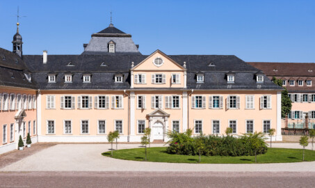 Schloss und Schlossgarten Schwetzingen - Schloss - Nordflügel / Hofkavaliersflügel - Von Süden - Blick vom Obergeschoss des Südflügels nach Norden auf den Ehrenhof, auf die Südfassade des Längsbaus (Mitte; heute genutzt vom Finanzamt) sowie auf die Ostfassade des Querbaus bzw. der Schlosskapelle (links); links der Mitte im Hintergrund das Dach des Oberen Wasserwerks (NordFlügel: L-förmig, Länge x Breite Längsbau ca. 50 x 14 Meter, Querbau ca. 15 x 12 Meter; Architekt: Johann Adam Breunig (1660-1727); Fertigstellung: 1710) (aufgenommen im August 2024, um die Mittagszeit)