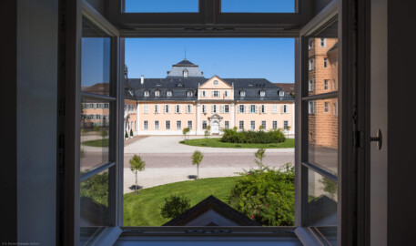 Schloss und Schlossgarten Schwetzingen - Schloss - Nordflügel / Hofkavaliersflügel - Von Süden - Blick durch das zentrale Fenster im Obergeschoss des Südflügels nach Norden auf den Ehrenhof, auf die Südfassade des Längsbaus (Mitte; heute genutzt vom Finanzamt) sowie auf die Ostfassade des Querbaus bzw. der Schlosskapelle (links); links oberhalb der Bildmitte im Hintergrund das Dach des Oberen Wasserwerks (NordFlügel: L-förmig, Länge x Breite Längsbau ca. 50 x 14 Meter, Querbau ca. 15 x 12 Meter; Architekt: Johann Adam Breunig (1660-1727); Fertigstellung: 1710) (aufgenommen im August 2024, um die Mittagszeit)