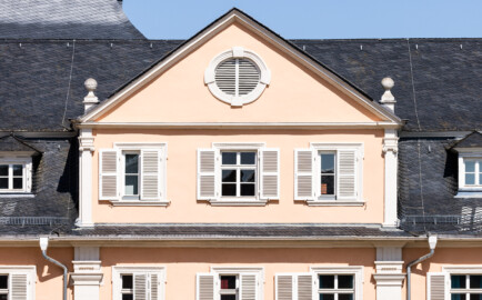 Schloss und Schlossgarten Schwetzingen - Schloss - Nordflügel / Hofkavaliersflügel - Längsbau - Mittelrisalit - Von Süden - Blick vom Obergeschoss des Südflügels nach Norden auf das Giebelfeld mit dem Ochsenauge und auf das Dachgeschoss des Mittelrisalits des Nordflügels; links der Mitte im Hintergrund das Dach des Oberen Wasserwerks (NordFlügel: L-förmig, Länge x Breite Längsbau ca. 50 x 14 Meter, Querbau ca. 15 x 12 Meter; Architekt: Johann Adam Breunig (1660-1727); Fertigstellung: 1710) (aufgenommen im August 2024, um die Mittagszeit)