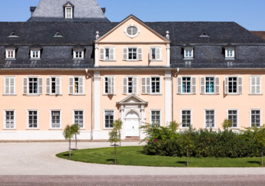 Schloss und Schlossgarten Schwetzingen - Schloss - Nordflügel / Hofkavaliersflügel - Längsbau - Von Süden - Blick vom Obergeschoss des Südflügels nach Norden auf den Ehrenhof und auf die Südfassade des Nordflügels; links der Mitte im Hintergrund das Dach des Oberen Wasserwerks (Nordflügel: L-förmig, Länge x Breite Längsbau ca. 50 x 14 Meter, Querbau ca. 15 x 12 Meter; Architekt: Johann Adam Breunig (1660-1727); Fertigstellung: 1710) (aufgenommen im August 2024, am frühen Nachmittag)