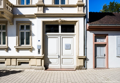 Schloss und Schlossgarten Schwetzingen - Schloss - Hochschule für Rechtspflege & Justizakademie - Verwaltungsbau - Von Osten - Blick von der Karlsruher Straße nach Westen auf den Eingangsbereich / das Eingangstor des Verwaltungsbaus; oben am Torrahmen aus Sandstein die Inschrift "1870"; rechts ein Teil der Ostfassade des Dozentenhauses (Verwaltungsbau: Länge x Breite ca. 15 x 15 Meter; letzte Renovierung: 2009-2012) (aufgenommen im August 2024, am späten Vormittag) Schloss und Schlossgarten Schwetzingen - Schloss - Hochschule für Rechtspflege & Justizakademie - Verwaltungsbau - Von Osten - Blick von der Karlsruher Straße nach Westen auf den Eingangsbereich / das Eingangstor des Verwaltungsbaus; oben am Torrahmen aus Sandstein die Inschrift "1870"; rechts ein Teil der Ostfassade des Dozentenhauses (Verwaltungsbau: Länge x Breite ca. 15 x 15 Meter; letzte Renovierung: 2009-2012) (aufgenommen im August 2024, am späten Vormittag)