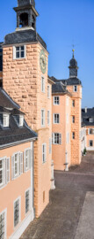 Schloss und Schlossgarten Schwetzingen - Schloss - Mittelbau - Hofseite - Von Südost - Blick vom Dachgeschoss des Südflügels nach Nordwesten auf den Ehrenhof und den Mittelbau; v.l.n.r.: Querbau des Südflügels, Südturm mit Uhr und Glocke, nördliches Treppenhaus, Nordturm, Querbau des Nordflügels mit der Schlosskapelle und Längsbau des Nordflügels (des Hofkavaliersflügels, heute vom Finanzamt genutzt) (Mittelbau: Länge Westseite ca. 45 Meter, Breite Süd-/Nordseite ca. 38 Meter; Wiederaufbau / Erweiterung: 1698-1717; Architekt: Johann Adam Breunig) (aufgenommen im August 2024, am Vormittag)