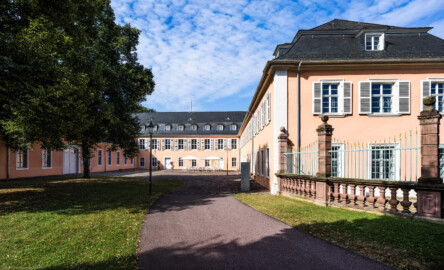 Schloss und Schlossgarten Schwetzingen - Schloss - Hof zwischen Südflügel und Küchenflügel - Von Osten - Blick vom Hofeingang nach Westen in den Hof, auf die Ostfassade des Südflügels (rechts), auf die Ostfassade des Querbaus des Küchenflügels (Mitte) sowie auf die Nordfassade des Längsbaus des Küchenbaus (links); ganz rechts ein Ende des Gitterzauns um den Ehrenhof des Schlosses (Südflügel: L-förmig, Länge x Breite Längsbau ca. 50 x 14 Meter, Querbau ca. 15 x 11 Meter; Architekt: Johann Adam Breunig (1660-1727); Fertigstellung: 1710; Küchenflügel: L-förmig, Länge x Breite Längsbau ca. 75 x 13 Meter, Querbau ca. 25 x 11 Meter; Architekt: Nicolas de Pigage (1723-1796); Fertigstellung: 1761; letzte Renovierung beider Flügel: 2009-2012) (aufgenommen im September 2024, am späten Vormittag)